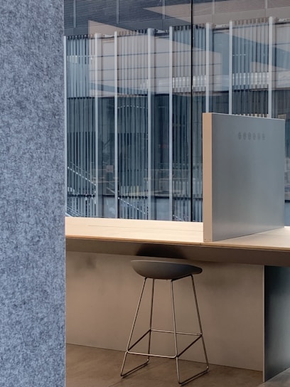 A modern office setup featuring a sleek stool placed under a light wooden desk. The backdrop includes large glass windows that reveal a view of an adjacent building's architecture with vertical lines. The interior design incorporates minimalist elements with a focus on clean lines and open space.