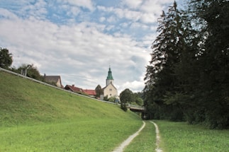 A scenic rural landscape featuring a church with a distinct green spire in the background. The image includes a grass-covered hill on the left and a pathway leading towards the church. Tall trees border the right side, and a road and some houses are visible in the distance under a partly cloudy sky.