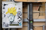 A utility box is decorated with street art stickers, prominently featuring a black-and-white portrait of a person with exaggerated yellow spiky hair. Various other stickers surround the portrait, including one with colorful abstract designs. The box is mounted on a metal pole, which is secured with a chain and padlock.