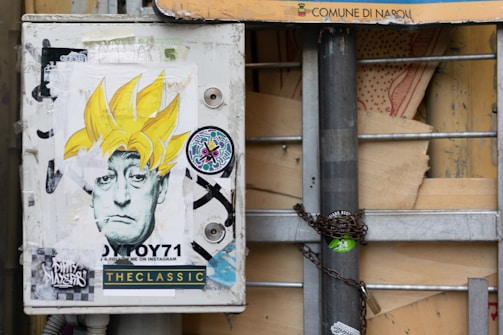 A utility box is decorated with street art stickers, prominently featuring a black-and-white portrait of a person with exaggerated yellow spiky hair. Various other stickers surround the portrait, including one with colorful abstract designs. The box is mounted on a metal pole, which is secured with a chain and padlock.