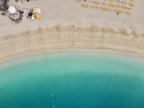 An aerial view of a serene beach with turquoise waters gently meeting the sandy shore. On the sand, bright yellow sun loungers are neatly lined up, and at the top left corner, a small beach cafe with colorful umbrellas and tables is visible.