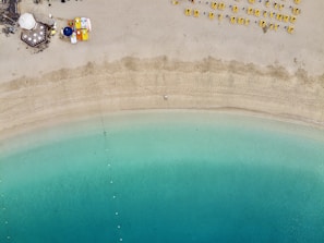 An aerial view of a serene beach with turquoise waters gently meeting the sandy shore. On the sand, bright yellow sun loungers are neatly lined up, and at the top left corner, a small beach cafe with colorful umbrellas and tables is visible.