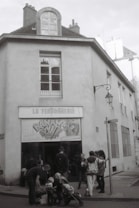 A black and white photograph of a building with a sign above the entrance reading 'La Ferronnerie'. Several people, including adults and children, are gathered in front of the entrance, some with strollers. The street corner has a vintage lantern and there are tall windows on the building's facade.