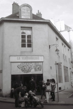 A black and white photograph of a building with a sign above the entrance reading 'La Ferronnerie'. Several people, including adults and children, are gathered in front of the entrance, some with strollers. The street corner has a vintage lantern and there are tall windows on the building's facade.