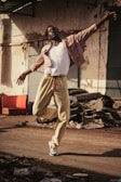 A person dances energetically on a dusty street, wearing a white tank top, light pink jacket, and beige pants. Debris and old car parts are scattered around, creating an industrial backdrop.