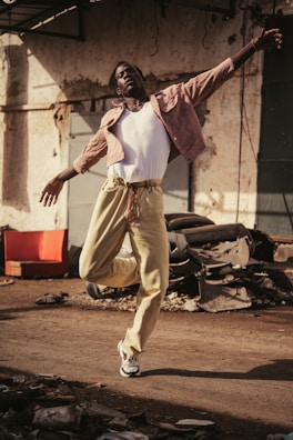 A person dances energetically on a dusty street, wearing a white tank top, light pink jacket, and beige pants. Debris and old car parts are scattered around, creating an industrial backdrop.