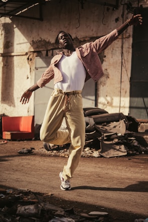 A person dances energetically on a dusty street, wearing a white tank top, light pink jacket, and beige pants. Debris and old car parts are scattered around, creating an industrial backdrop.