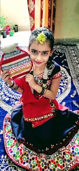 A child dressed in colorful traditional attire sits on a patterned textile, holding a decorative object. Her hair is adorned with a yellow flower, and she has painted designs on her face. The background includes a curtain and several small potted plants.