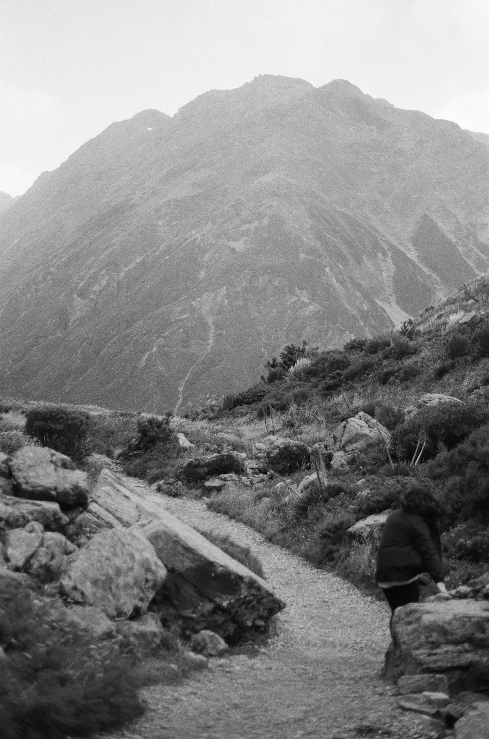 grayscale photo of person sitting on rock near mountain