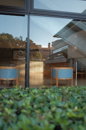 A modern architectural interior is seen through a window. There are two minimalist wooden benches with light blue cushions. The window reflects a partially visible urban skyline and trees. The interior features large open spaces with a glossy floor and contemporary design elements.