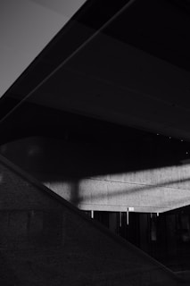 A minimalistic black and white composition with strong geometric lines and shadows. The image features intersecting architectural elements with harsh light casting distinct shadows on textured concrete surfaces.