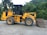 A yellow front loader vehicle with large tires is parked on a gravel surface, surrounded by dense green foliage and hills in the background. The machine has a prominent logo on the side, indicating the brand 'Liugong'.