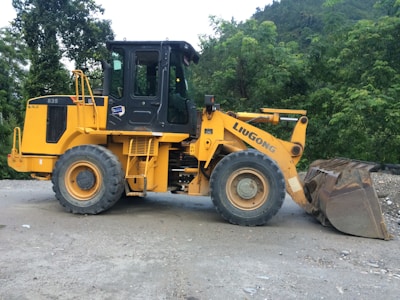 A yellow front loader vehicle with large tires is parked on a gravel surface, surrounded by dense green foliage and hills in the background. The machine has a prominent logo on the side, indicating the brand 'Liugong'.