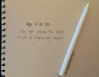 A spiral-bound notebook with a handwritten quote about the ages 15 to 23 and the temporary nature of people encountered during that time. A white pen is placed next to the text on the right side.