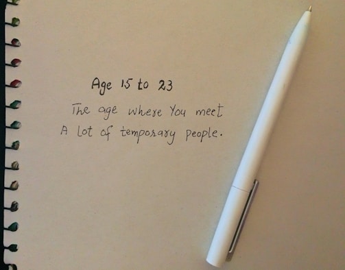 A spiral-bound notebook with a handwritten quote about the ages 15 to 23 and the temporary nature of people encountered during that time. A white pen is placed next to the text on the right side.