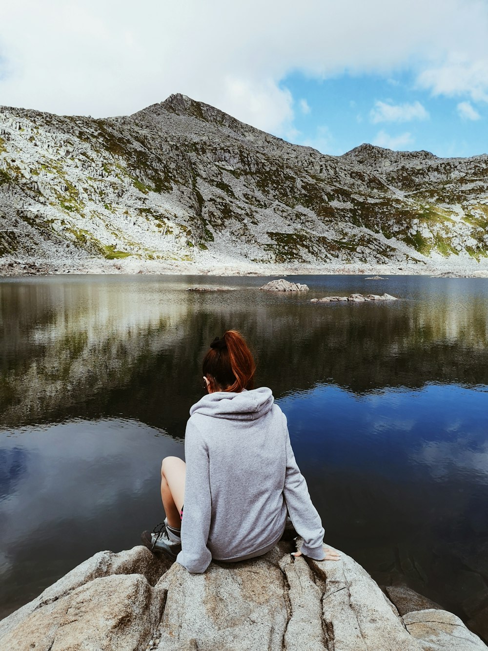 woman in gray hoodie sitting on rock near lake during daytime