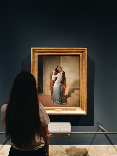 A person is observing a framed painting displayed on a museum wall. The painting depicts a romantic scene of two people embracing and kissing on a stone staircase. The woman has long hair, and the man wears a hat. The colors in the painting are warm and vivid, contrasting with the dark surroundings.