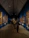 A gallery with several paintings adorning the walls. The ceiling is high and triangular, featuring architectural lighting. Visitors are observing the paintings, some standing close to the artworks. The floor has a polished look, and the walls are painted a deep blue, creating a vibrant backdrop for the framed artworks.
