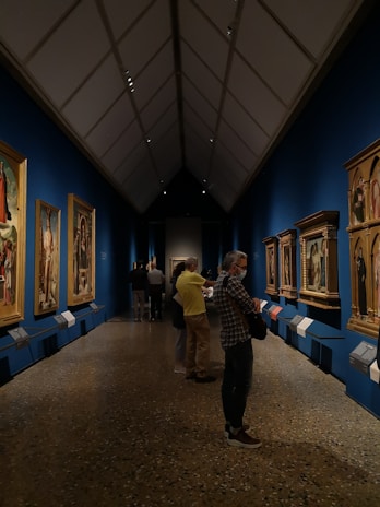 A gallery with several paintings adorning the walls. The ceiling is high and triangular, featuring architectural lighting. Visitors are observing the paintings, some standing close to the artworks. The floor has a polished look, and the walls are painted a deep blue, creating a vibrant backdrop for the framed artworks.