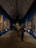 A gallery with several paintings adorning the walls. The ceiling is high and triangular, featuring architectural lighting. Visitors are observing the paintings, some standing close to the artworks. The floor has a polished look, and the walls are painted a deep blue, creating a vibrant backdrop for the framed artworks.