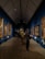 A gallery with several paintings adorning the walls. The ceiling is high and triangular, featuring architectural lighting. Visitors are observing the paintings, some standing close to the artworks. The floor has a polished look, and the walls are painted a deep blue, creating a vibrant backdrop for the framed artworks.