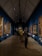 A gallery with several paintings adorning the walls. The ceiling is high and triangular, featuring architectural lighting. Visitors are observing the paintings, some standing close to the artworks. The floor has a polished look, and the walls are painted a deep blue, creating a vibrant backdrop for the framed artworks.