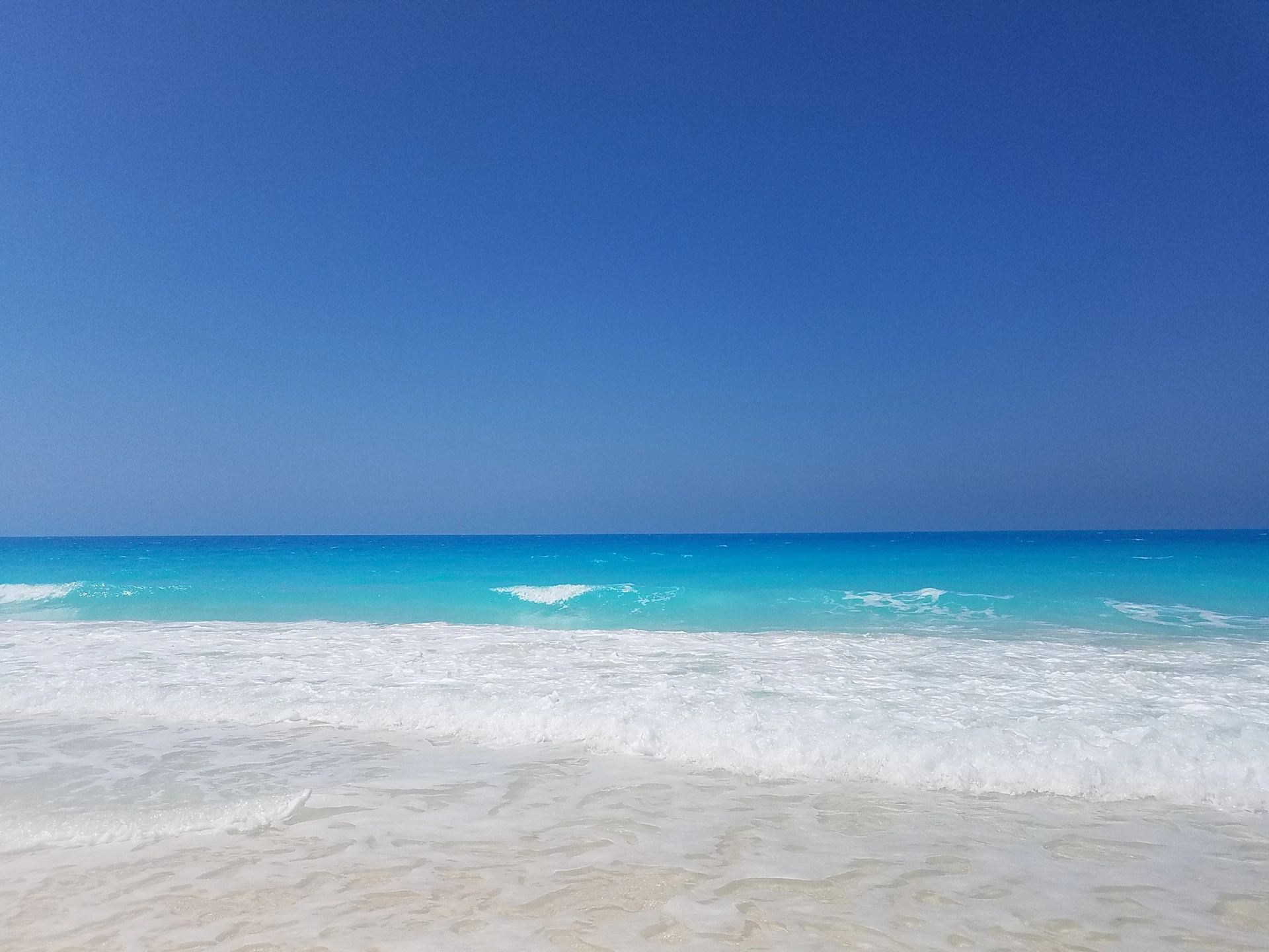 A tranquil beach with crystal-clear turquoise water, gentle waves, and smooth white sand under a peaceful blue sky.