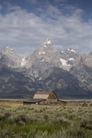 A rustic wooden barn sits in the foreground of a vast, open field with sagebrush. Towering, snow-capped mountain peaks rise majestically in the background under a partly cloudy sky.