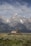 A rustic wooden barn sits in the foreground of a vast, open field with sagebrush. Towering, snow-capped mountain peaks rise majestically in the background under a partly cloudy sky.