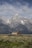 A rustic wooden barn sits in the foreground of a vast, open field with sagebrush. Towering, snow-capped mountain peaks rise majestically in the background under a partly cloudy sky.