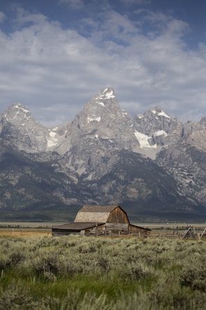 A rustic wooden barn sits in the foreground of a vast, open field with sagebrush. Towering, snow-capped mountain peaks rise majestically in the background under a partly cloudy sky.