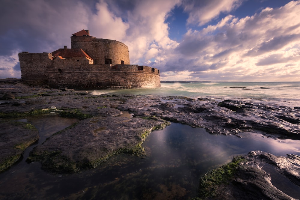 brown concrete building on top of rocky hill by the sea under blue and white cloudy