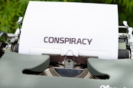 A vintage typewriter with a sheet of paper inserted, displaying the word 'CONSPIRACY' in bold, uppercase letters. The typewriter is set against a blurred green background.