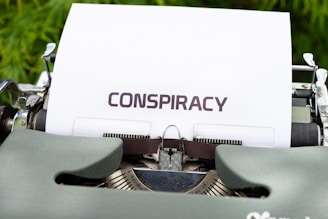 A vintage typewriter with a sheet of paper inserted, displaying the word 'CONSPIRACY' in bold, uppercase letters. The typewriter is set against a blurred green background.