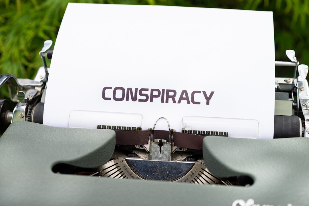 Parte de cima de máquina de escrever com gramado ao fundo onde se vê papel saindo com a palavra “Conspiracy”.