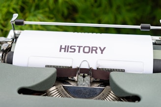 A close-up view of a typewriter with a sheet of paper in it displaying the word 'HISTORY'. The typewriter has a vintage design with visible keys and mechanical parts. The background features a blurred green foliage, adding a natural element to the scene.