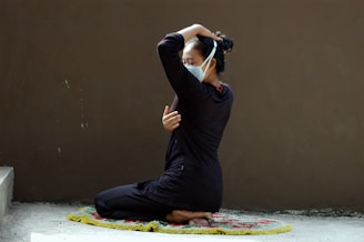 A person wearing a face mask is sitting on a colorful mat in a yoga pose with one arm bent over their head and the other arm on their shoulder. The background is a plain, neutral-colored wall.
