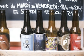A row of six beer bottles with various unique and colorful labels is arranged on a wooden shelf. Behind them, a chalkboard contains handwritten text in French. The scene is likely set in a casual environment like a bar or caf&eacute;.