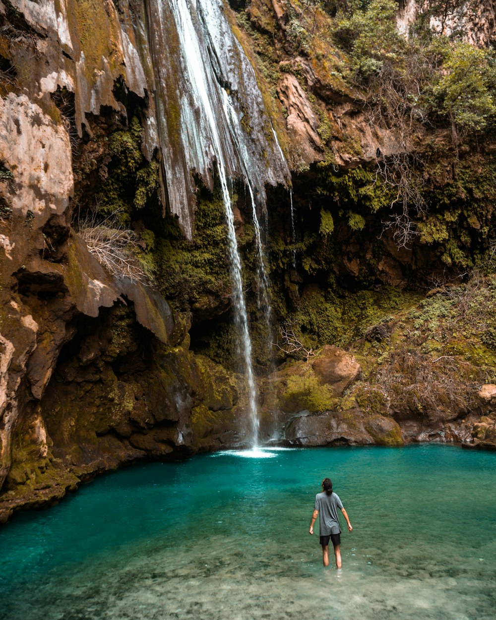 uomo in camicia nera e jeans blu denim in piedi sulle cascate d'acqua durante il giorno