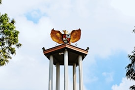 A tall structure features a golden eagle with outstretched wings perched on top. The sculpture is adorned with a shield displaying symbols, resting on a high platform supported by columns. The structure is surrounded by greenery, with clear blue skies in the background.