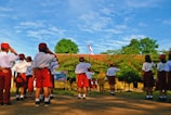 A group of children wearing uniforms consisting of white shirts, red shorts, and red caps are saluting a flag in an outdoor setting. The background features a lush green landscape with trees and a building with a red roof. The sky is clear with scattered clouds, indicating a sunny day.