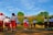 A group of children wearing uniforms consisting of white shirts, red shorts, and red caps are saluting a flag in an outdoor setting. The background features a lush green landscape with trees and a building with a red roof. The sky is clear with scattered clouds, indicating a sunny day.