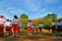A group of children wearing uniforms consisting of white shirts, red shorts, and red caps are saluting a flag in an outdoor setting. The background features a lush green landscape with trees and a building with a red roof. The sky is clear with scattered clouds, indicating a sunny day.