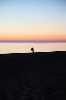 A serene beach scene during twilight where a couple stands silhouetted against a pastel-colored sky and calm sea, evoking a sense of tranquility and romance.