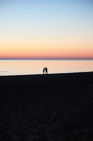 A serene beach scene during twilight where a couple stands silhouetted against a pastel-colored sky and calm sea, evoking a sense of tranquility and romance.