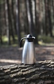 A metal thermos with a black handle and lid rests on a tree log in a forest setting. Tall trees with blurred trunks and sunlight filtering through create a natural, serene environment.