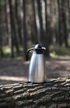 A metal thermos with a black handle and lid rests on a tree log in a forest setting. Tall trees with blurred trunks and sunlight filtering through create a natural, serene environment.