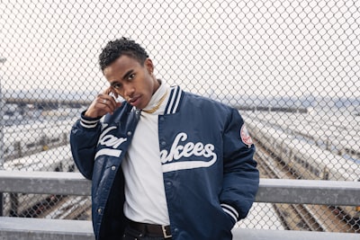 man in blue and white adidas zip up jacket standing beside gray metal fence during daytime yankees google meet background