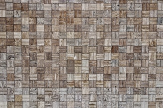 A close-up view of a textured wall composed of small rectangular stone tiles arranged in a grid pattern. The colors of the tiles vary, featuring different shades of brown, beige, and gray. The surface shows natural stone patterns with slight variations in texture and appearance.