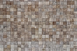 A close-up view of a textured wall composed of small rectangular stone tiles arranged in a grid pattern. The colors of the tiles vary, featuring different shades of brown, beige, and gray. The surface shows natural stone patterns with slight variations in texture and appearance.